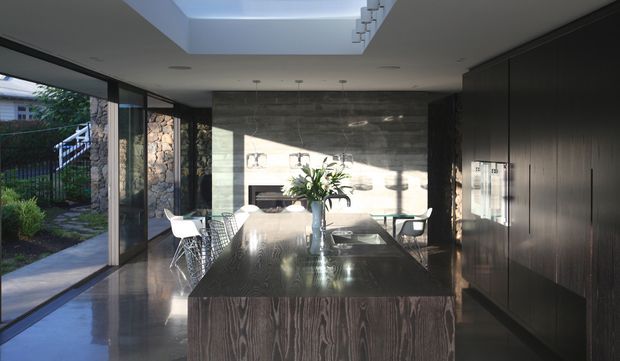 A contemporary open-plan kitchen and dining space with a large dark timber island, integrated dark cabinetry, polished concrete or stone flooring, and a dark stone feature wall surrounding a fireplace. Full-height glazing opens to a landscaped exterior, while a roof skylight and linear pendant lighting bring in natural light. A dining table and chairs sit beside the island.