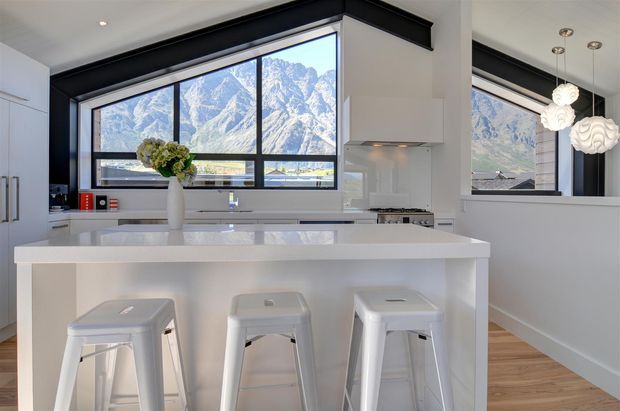Bright contemporary white kitchen with a large central island and three metal bar stools. Black-framed picture windows provide dramatic mountain views, while white cabinetry, a glossy light benchtop, timber flooring, and sculptural pendant lights create a clean, refined interior.