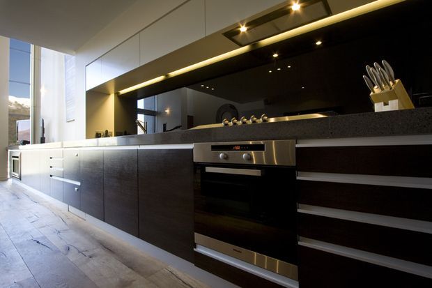 A sleek contemporary galley-style kitchen with a long run of dark flat-panel cabinetry, integrated stainless-steel oven, black stone-look benchtop, and a glossy black splashback or mirrored wall. Under-cabinet and recessed strip lighting illuminate the work zone. Pale timber flooring and tall glazing at the end of the room add contrast and natural light.
