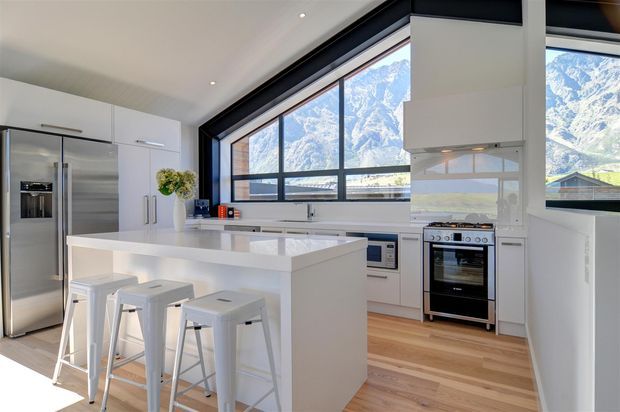 Bright contemporary kitchen with white full-height cabinetry, a large white island with seating for three, stainless-steel appliances, pale timber flooring, and expansive dark-framed windows overlooking snow-capped mountains.