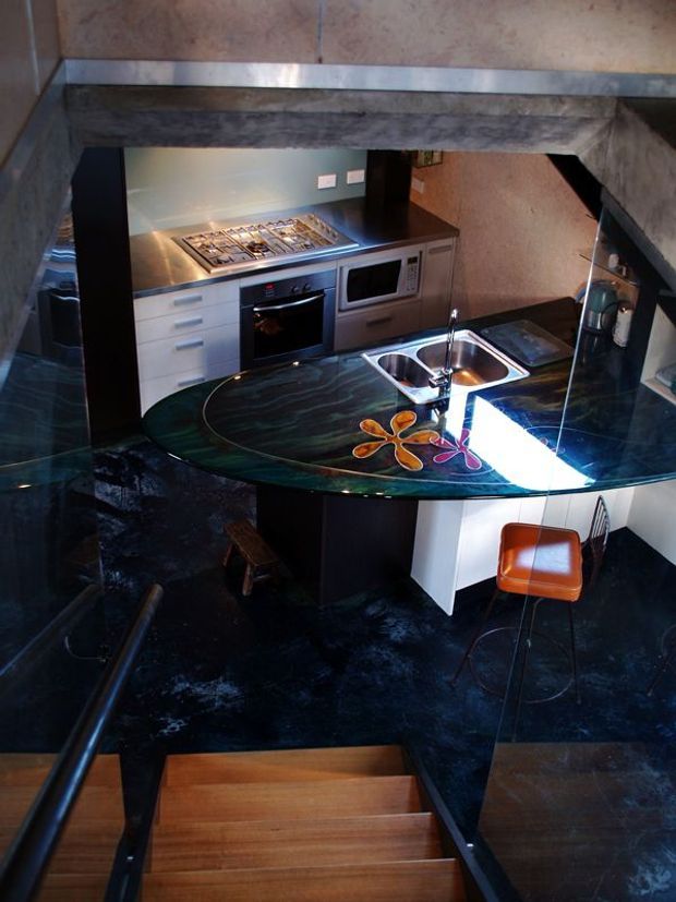 View from a staircase into a compact, highly styled kitchen with a curved peninsula island. The kitchen includes stainless-steel appliances, white drawers, dark cabinetry, a black veined stone-looking benchtop, integrated double sink, glass-like curved breakfast bar, and colourful floral inlay detail. Dark polished flooring, timber stair treads, and a warm orange bar stool create a dramatic contemporary interior.