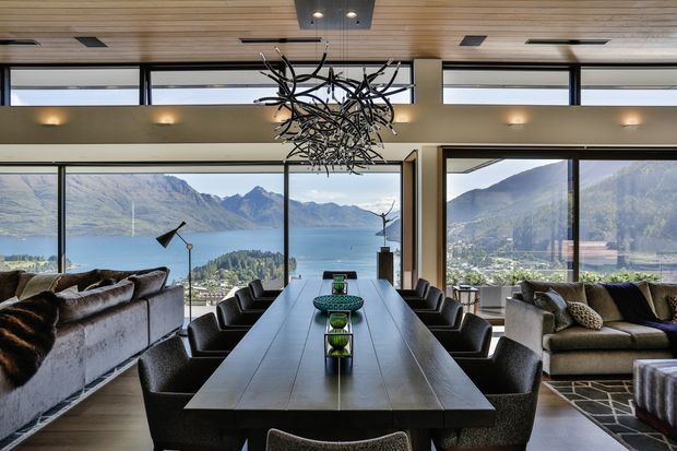 A refined open-plan dining and living area with a long dark dining table, multiple upholstered dining chairs, sofas, and a dramatic sculptural chandelier. Floor-to-ceiling glazing frames expansive mountain and lake views, while a timber-lined ceiling, dark-framed windows, and warm neutral finishes create a contemporary alpine atmosphere.