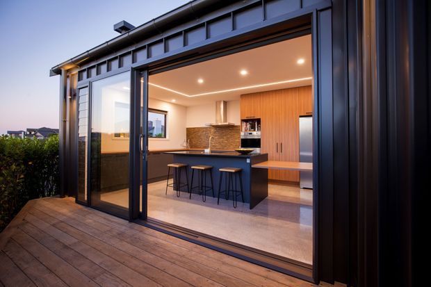 A contemporary kitchen opening onto a timber deck through large black-framed sliding glass doors. The kitchen features a central island with seating for four, warm timber cabinetry, a dark benchtop, integrated appliances, recessed ceiling lighting, and a textured stone-look splashback. Dark vertical exterior cladding and structural framing surround the opening.