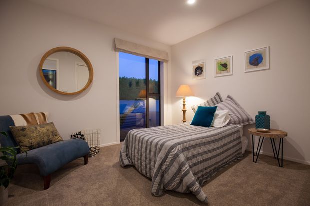 A cozy, softly lit bedroom with a single bed dressed in a grey-and-white striped coverlet, layered pillows, and a teal accent cushion. The room includes a blue upholstered armchair, round timber-framed wall mirror, bedside table with lamp, framed artwork, carpeted flooring, and a glazed door or window opening to an exterior balcony or terrace.