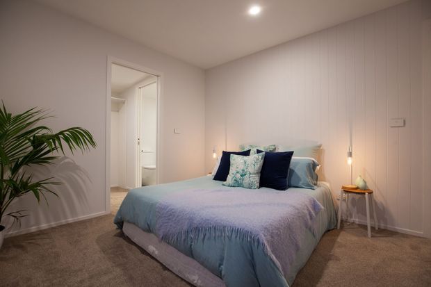 A softly lit bedroom with a low-profile upholstered bed dressed in pale blue and lavender bedding, dark navy and patterned cushions, and warm pendant bedside lights. The room has light neutral walls, carpeted flooring, white trim and doors, a small bedside table, and a leafy indoor plant. An open doorway provides a view into an adjacent bathroom or hallway.