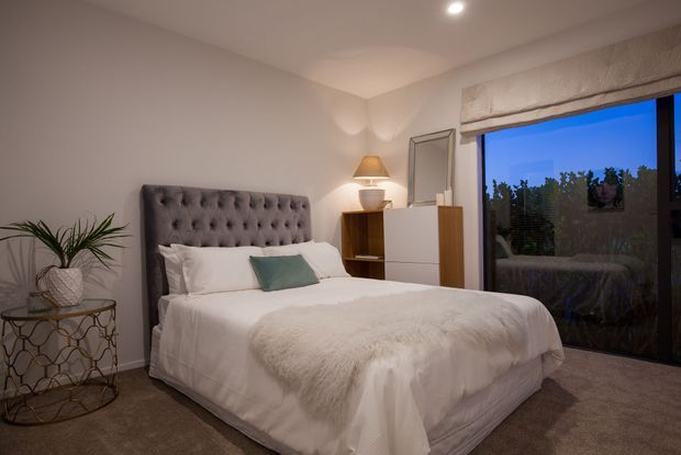 A softly lit, contemporary bedroom with a large upholstered button-tufted bed, white bedding, a pale teal accent cushion, and a textured throw. A timber and white bedside/storage unit with a table lamp and mirror sits beside the bed. The room has carpeted flooring, neutral walls, a potted plant on a metal side table, and a large glazed opening with a Roman blind showing an evening exterior.