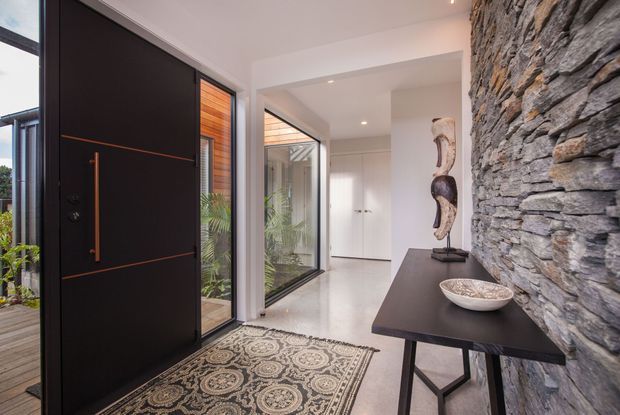 A refined contemporary entryway with a tall matte-black pivot door featuring warm timber detailing, extensive glazing, and a patterned rug. A dramatic stacked-stone feature wall runs along the right side, paired with a slim dark console and sculptural decor. White walls, recessed lighting, and indoor greenery create a bright, polished entrance.