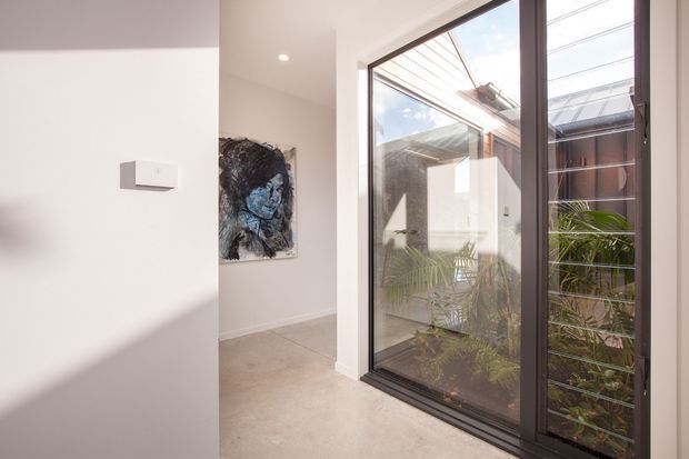Bright minimalist interior hallway or entry area with polished concrete flooring, white walls, and large dark-framed glazed doors opening onto a planted courtyard. A monochrome artwork hangs in the passage, while sunlight creates strong geometric shadows across the walls and floor.