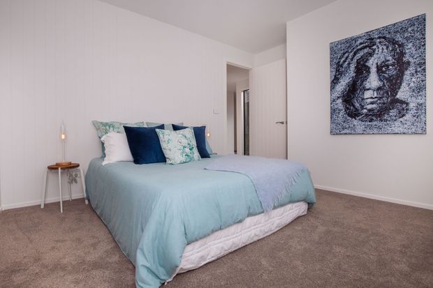 A softly styled bedroom with a large bed dressed in pale blue bedding and layered white, navy, and patterned cushions. The room has white walls and ceiling, wall-to-wall taupe carpet, a small bedside table with a pendant light, an open white door, and monochrome portrait artwork. The overall atmosphere is calm, airy, and understated.