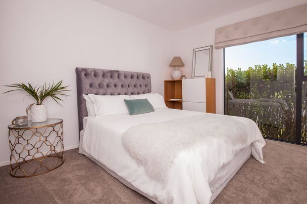 A bright, contemporary bedroom with a tufted upholstered bed, white bedding, a small muted blue accent cushion, and soft taupe carpeting. A round metal-and-glass side table with a potted plant sits beside the bed, while a timber and white built-in storage unit, table lamp, and large glazed door or window with a Roman blind occupy the far wall.