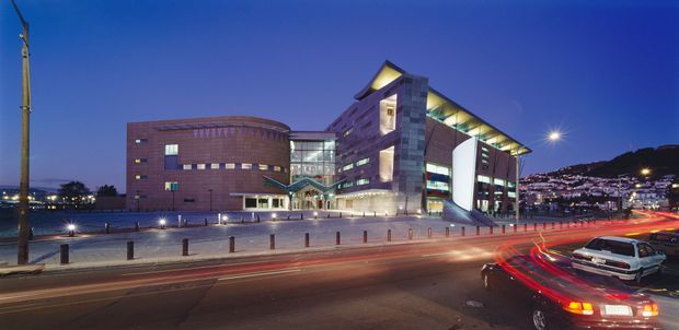 Night-time exterior view of a large contemporary multi-storey institutional or commercial building beside a busy urban roadway. The building has a distinctive curved brick-clad wing, a taller angular central volume with exposed structural expression, extensive glazed circulation areas, and illuminated interiors. Traffic light trails, street lighting, bollards, and a hilly urban landscape are visible in the surroundings.