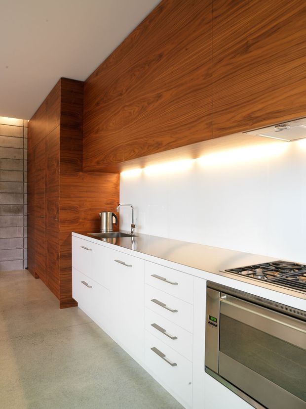 A contemporary galley kitchen with a long white benchtop and white handle-front cabinetry, integrated stainless-steel oven and gas cooktop, and a small undermount sink. Richly grained dark timber cabinetry and wall panelling form a strong feature surface above and beside the kitchen, while concealed under-cabinet lighting illuminates a plain white splashback. The space has a polished concrete floor and a narrow adjoining passage.