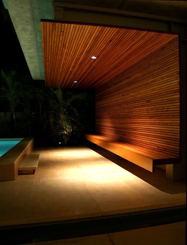 Night-time exterior poolside terrace featuring a dramatic cantilevered timber-slat canopy and matching vertical timber-slat feature wall. Warm recessed lighting washes over a long built-in bench and pale stone-paved terrace, with tropical planting and the edge of a swimming pool visible at left. The design has a refined, resort-like outdoor atmosphere with strong horizontal and vertical linear detailing.