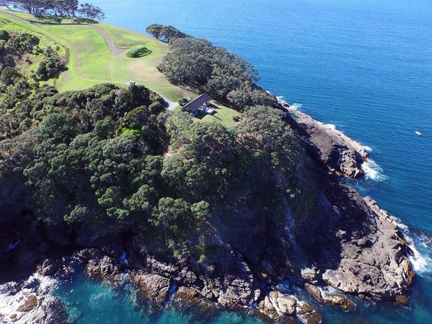 Aerial view of a heavily wooded coastal headland with rocky cliffs extending into clear blue ocean. A small building sits among trees near an open manicured lawn, with a winding driveway or pathway visible inland. The image primarily captures the dramatic waterfront landscape and rugged shoreline.