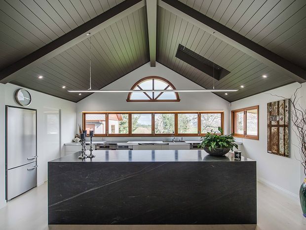 A bright contemporary kitchen centred around a large dark stone island with an integrated cooktop and seating-side work surface. The room has a dramatic pitched, timber-lined ceiling with dark exposed beams, white walls, extensive timber-framed windows, a high arched feature window, and minimalist white cabinetry. A linear pendant light, indoor plants, and restrained decorative details complete the space.