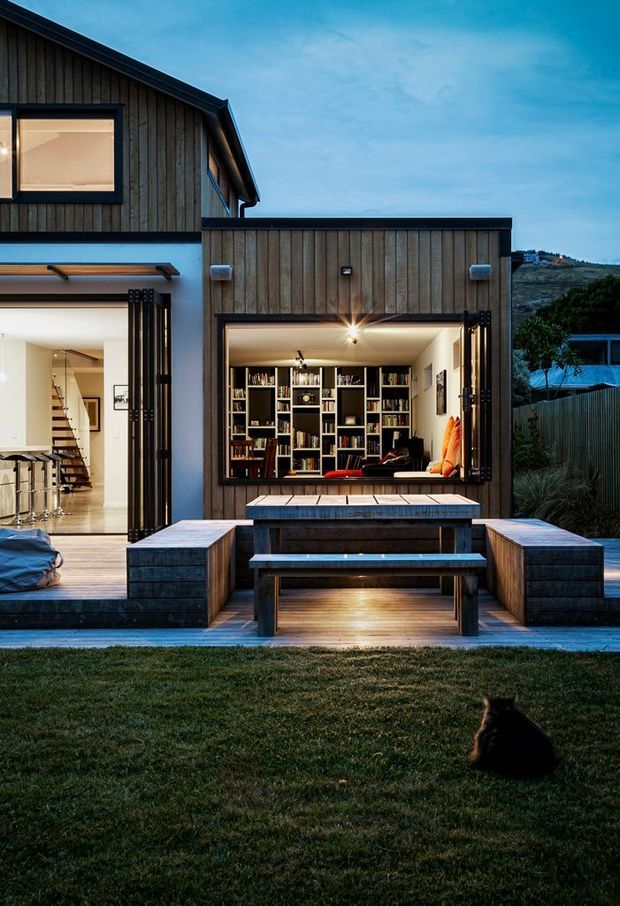 A contemporary two-storey residential exterior at dusk, clad in vertical dark timber boards with black-framed glazing and large bi-fold openings. The lower level opens onto a timber deck with a built-in outdoor dining bench and table, while a brightly lit interior library and living area are visible through the openings. A landscaped lawn occupies the foreground, with a sloping vegetated site beyond.