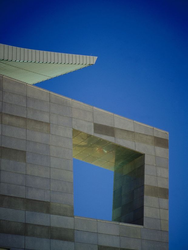 Upward-looking architectural photograph of a modern institutional or commercial facade clad in large rectangular grey panels. A prominent rectangular void cuts through the facade, with a reflective metallic soffit and deep reveals, while a projecting ribbed roof or canopy extends at the upper left. The composition emphasizes geometric form, sharp edges, material joints, and contrast against a clear deep-blue sky.