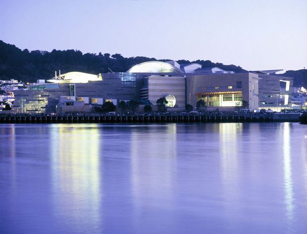 A wide dusk exterior view of a large contemporary waterfront cultural or commercial complex across calm water. The building has multiple interconnected volumes, curved and domed roof forms, extensive glazing, horizontal wall bands, and illuminated interiors. A wooded hillside and surrounding urban development are visible behind it, with strong purple-blue evening light reflected across the water.