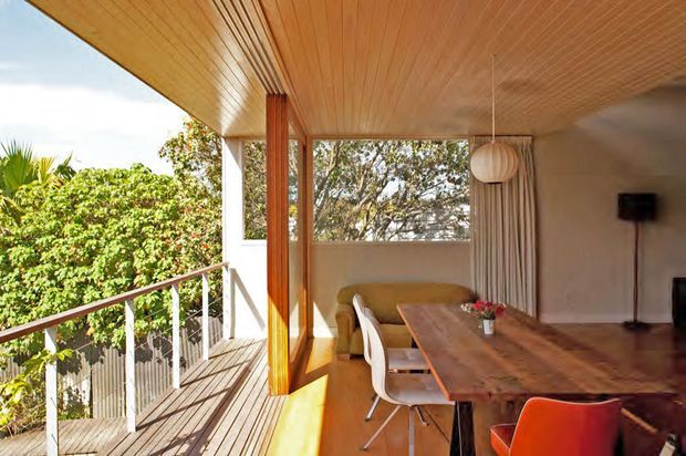 A warm, mid-century-inspired dining and living area opening onto a timber balcony. The room features a timber-lined ceiling, extensive glazing, slender timber posts, a long rustic timber dining table with modern chairs, a sculptural pendant light, and lush greenery visible outside.