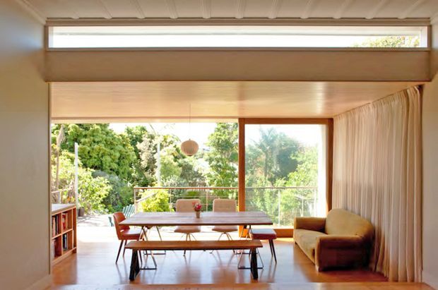 Warm mid-century-inspired dining and living area with a timber-lined ceiling, broad glazed opening to leafy greenery, a wooden dining table with bench seating, compact bookcase, armchair, pendant light, and full-height curtains. The space is bright and connected to the outdoors.
