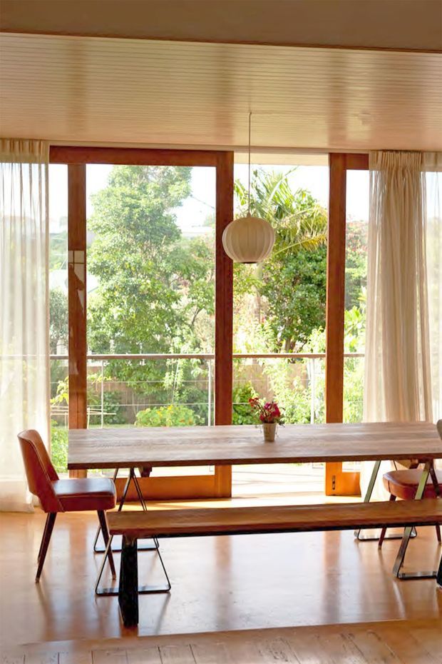 Bright dining room with a long timber dining table, matching wooden bench, and mid-century style reddish upholstered chairs. The room features expansive timber-framed glazing with sheer curtains, a light timber slatted ceiling, polished light flooring, and a spherical pendant light. Lush garden greenery is visible beyond the windows.
