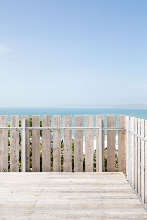A bright elevated timber deck or terrace enclosed by a vertical weathered-wood privacy balustrade, overlooking a turquoise ocean and distant coastline. The scene is minimalist and coastal, with pale timber decking, clear blue sky, and abundant natural light.