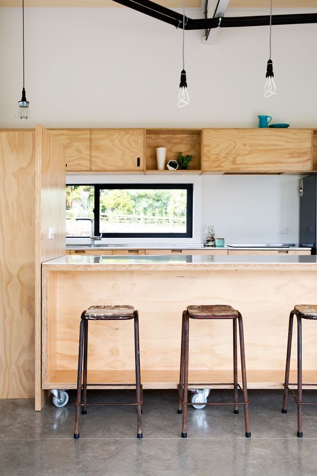 Bright contemporary kitchen featuring extensive light plywood cabinetry, a large island with a reflective benchtop, black-framed window, exposed ceiling services, pendant lights, and three industrial-style bar stools. The polished concrete floor and simple open shelving create a clean, functional aesthetic.