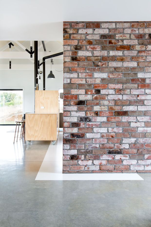Bright open-plan industrial interior featuring a large exposed red and weathered white brick feature wall, polished concrete flooring, exposed black structural framing, and simple plywood cabinetry or mobile storage. Large glazing brings in abundant daylight, with bar stools and pendant lighting visible in the workspace beyond.