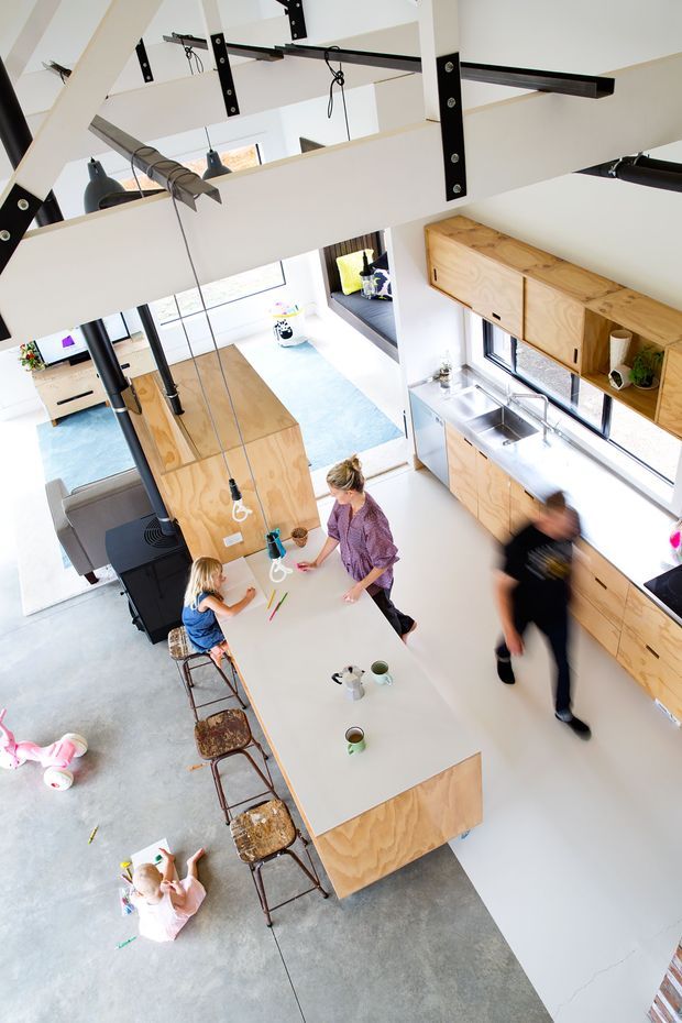 High-angle view of an open-plan kitchen and living area with a large plywood-clad island and matching timber cabinetry, white surfaces, polished concrete flooring, exposed black structural beams and hanging pendant lights. The space includes a dining/work island with stools, a lounge area, large windows, and several people using the room.