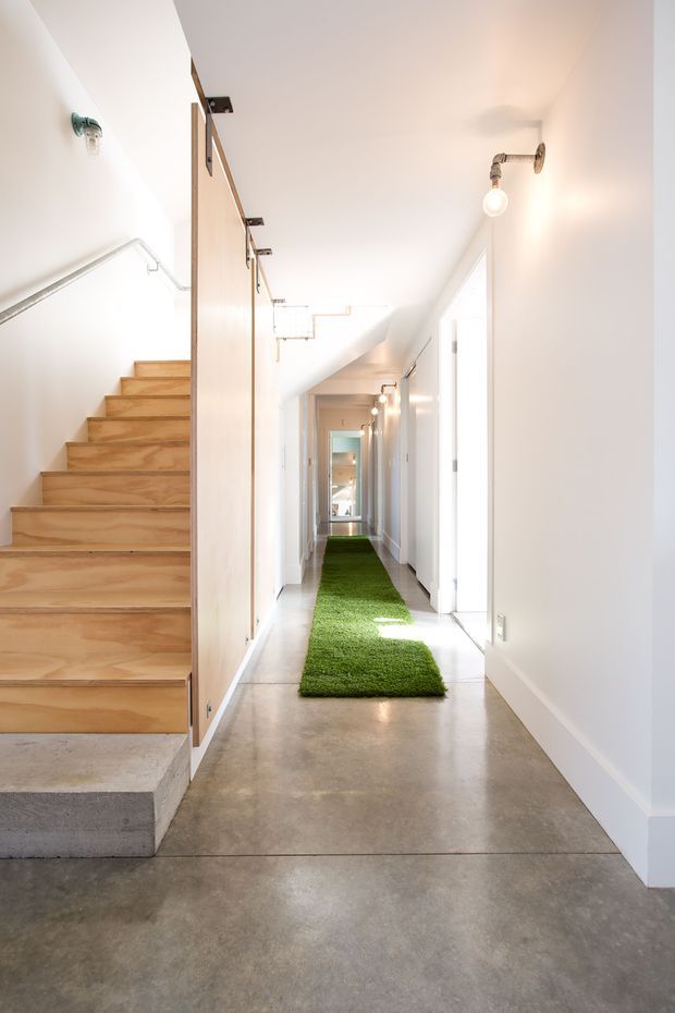 A bright, minimalist hallway with polished concrete flooring, white walls, and a long narrow circulation path. A timber staircase with a simple metal handrail rises on the left beside tall pale-wood storage or partition panels. White doorways line the corridor, which is illuminated by exposed wall-mounted and ceiling-mounted lights. A narrow green runner adds contrast along the floor.
