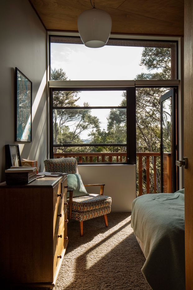 A warm, mid-century-inspired bedroom with a timber-lined ceiling, large dark-framed windows and a glazed door opening to a leafy balcony or terrace. The room contains a bed with muted green bedding, a patterned upholstered armchair, and a dark timber chest of drawers. Strong sunlight casts long shadows across the textured carpet, creating a calm connection to the surrounding trees.