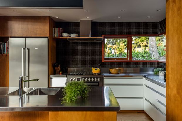 A contemporary kitchen with a dark tiled splashback, warm timber cabinetry and framing, white lower cabinets, stainless-steel appliances, and a large central island with an integrated sink and tap. A wide timber-framed window overlooks a leafy garden, while recessed ceiling lights provide illumination.