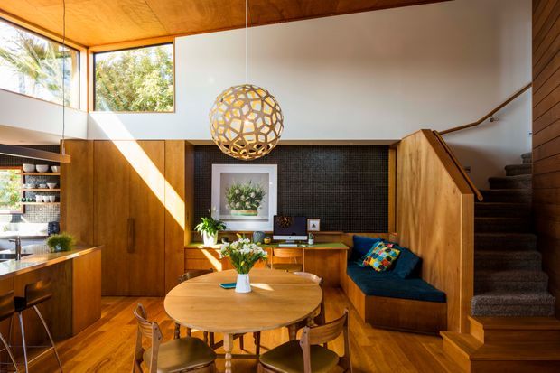 A warm, timber-lined open-plan interior combining a dining area, kitchen, study nook and staircase. The room features a round timber dining table with wooden chairs, timber cabinetry and flooring, a compact kitchen at left, a built-in desk beneath a dark textured feature wall, and a navy built-in window seat. High glazing and a partially raised ceiling bring in strong natural light, while a sculptural spherical pendant provides a focal point.