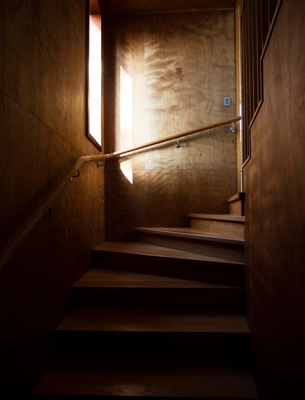 A narrow, enclosed staircase rising through a timber-lined stairwell. Dark stained plywood or timber panels cover the walls and ceiling, with matching wooden treads, simple timber handrails, and small metal brackets. A narrow vertical window introduces a strong shaft of natural light, creating a warm, dramatic and intimate atmosphere.