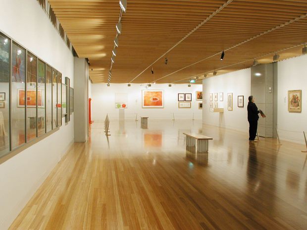 A spacious contemporary art gallery interior with polished timber flooring, white walls, framed artworks, display plinths, and a distinctive slatted timber ceiling with track lighting. The open exhibition space is viewed from inside, with a visitor standing near the artwork-covered walls.