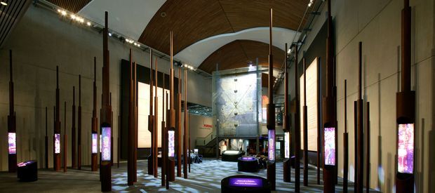 A dimly lit contemporary interior resembling an immersive exhibition or commercial gallery space. Tall slender timber or dark-wood vertical poles line both sides of a broad walkway, interspersed with illuminated digital display columns and low glowing plinths. Large warm backlit wall panels flank the space, while the ceiling combines exposed timber slats with a sweeping curved white architectural form. The far end features a large metallic or industrial wall installation. The atmosphere is dramatic, refined, and exhibition-focused.
