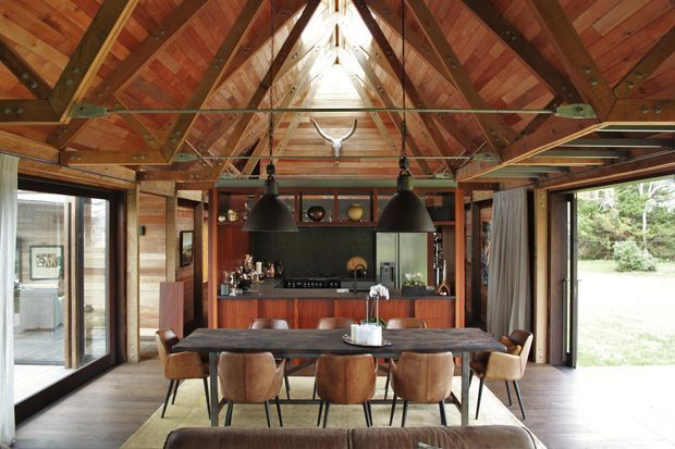 A warm, rustic open-plan dining and kitchen interior with a long timber dining table, leather dining chairs, dark pendant lights, and a compact kitchen with timber cabinetry and dark benchtops. The room has dramatic exposed timber roof framing, a high pitched ceiling with a central skylight or clerestory opening, extensive glazing, and direct views to greenery outside.