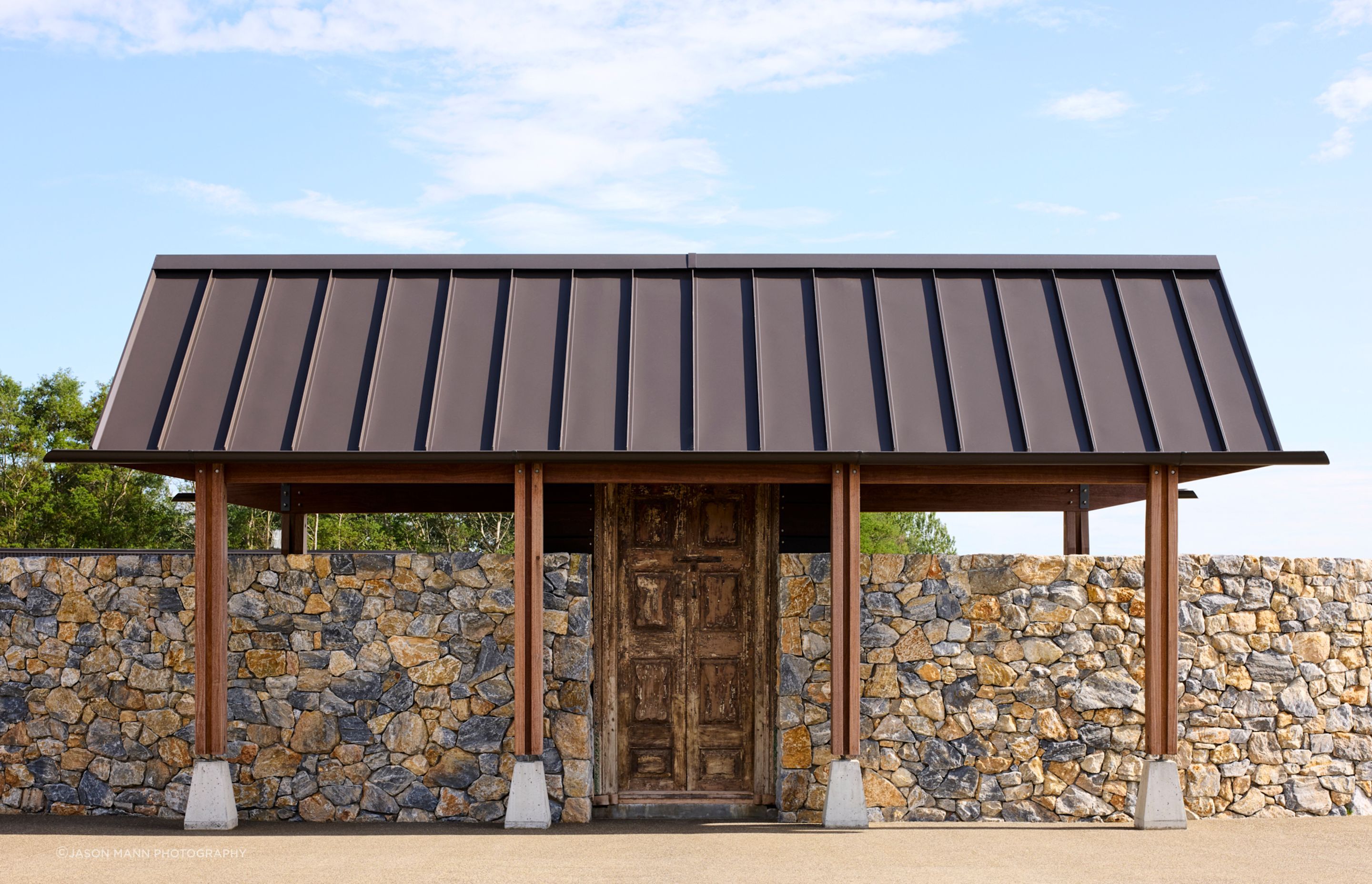 The beautiful, architecturally designed woodshed features a curved stone wall, with timber posts sitting on elegantly formed concrete plinths, holding up the roof.