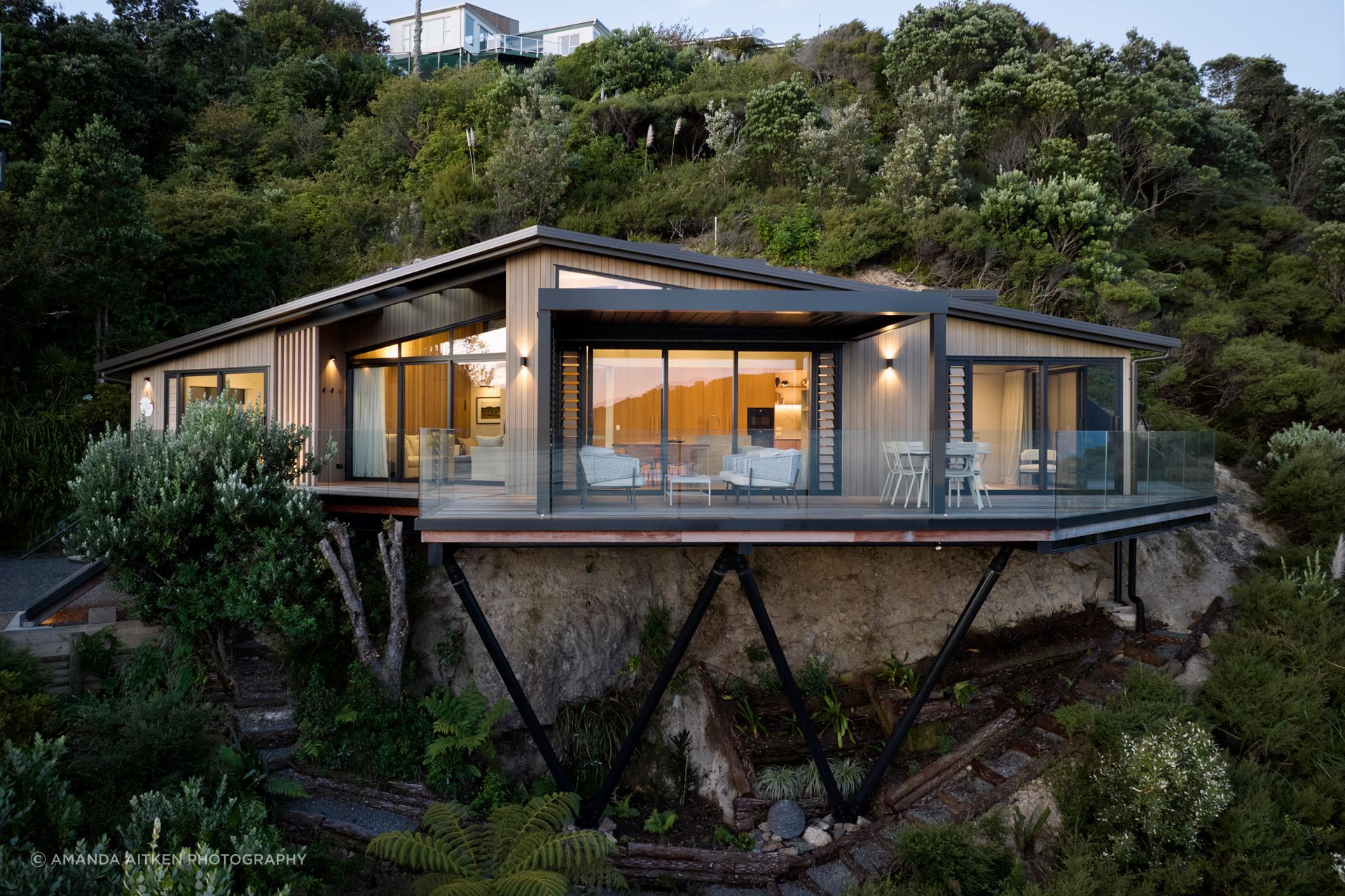 Nestled on the slopes of Mount Paku, Cliff Hanger House captures sweeping views of Tairua Harbour and Slipper Island, appearing to hover above the rugged coastline 35 metres below