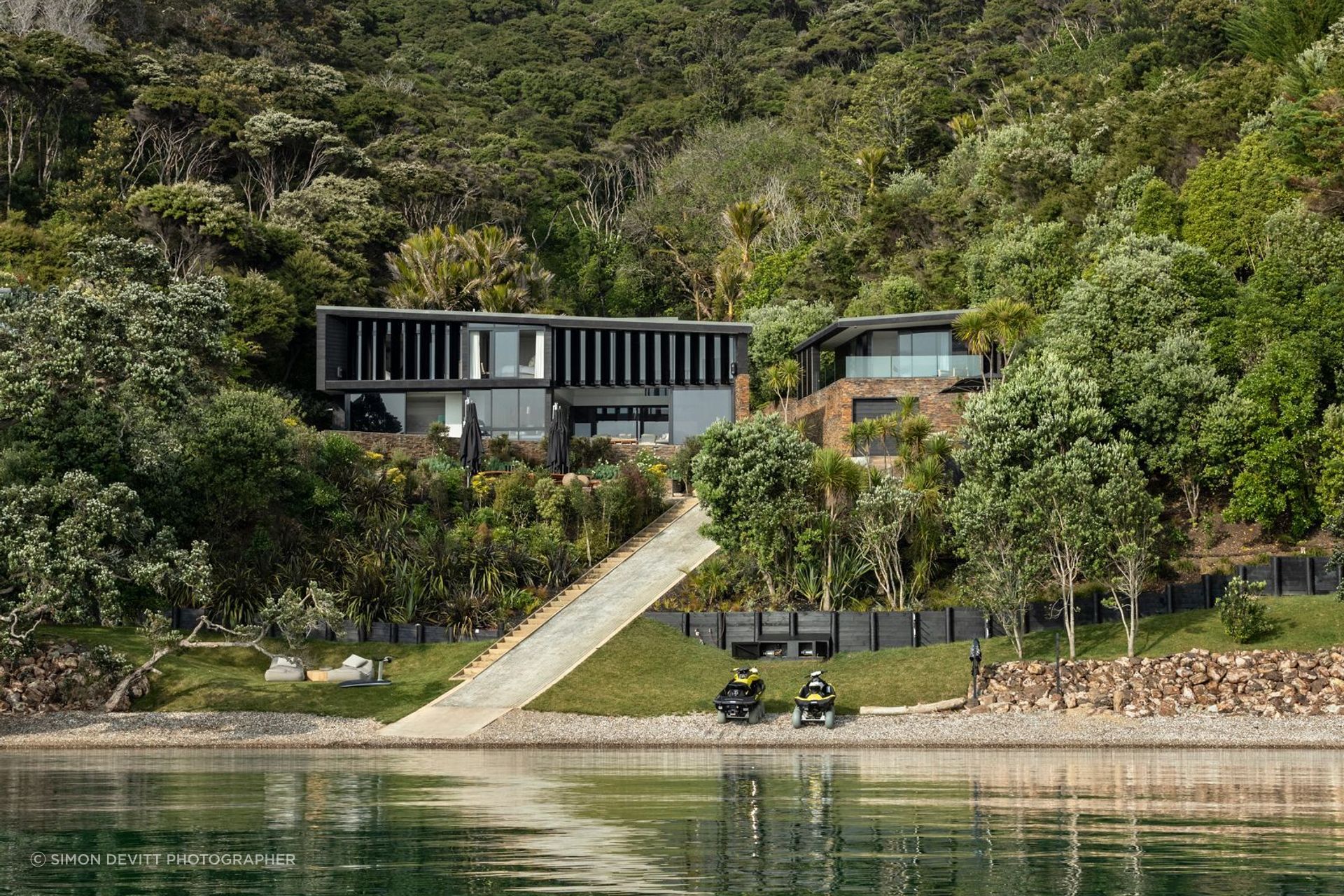 At Waikopua Bay, the house settles into the lowest part of the valley, its pavilions framed by existing nīkau and restored native bush rather than standing apart from them.