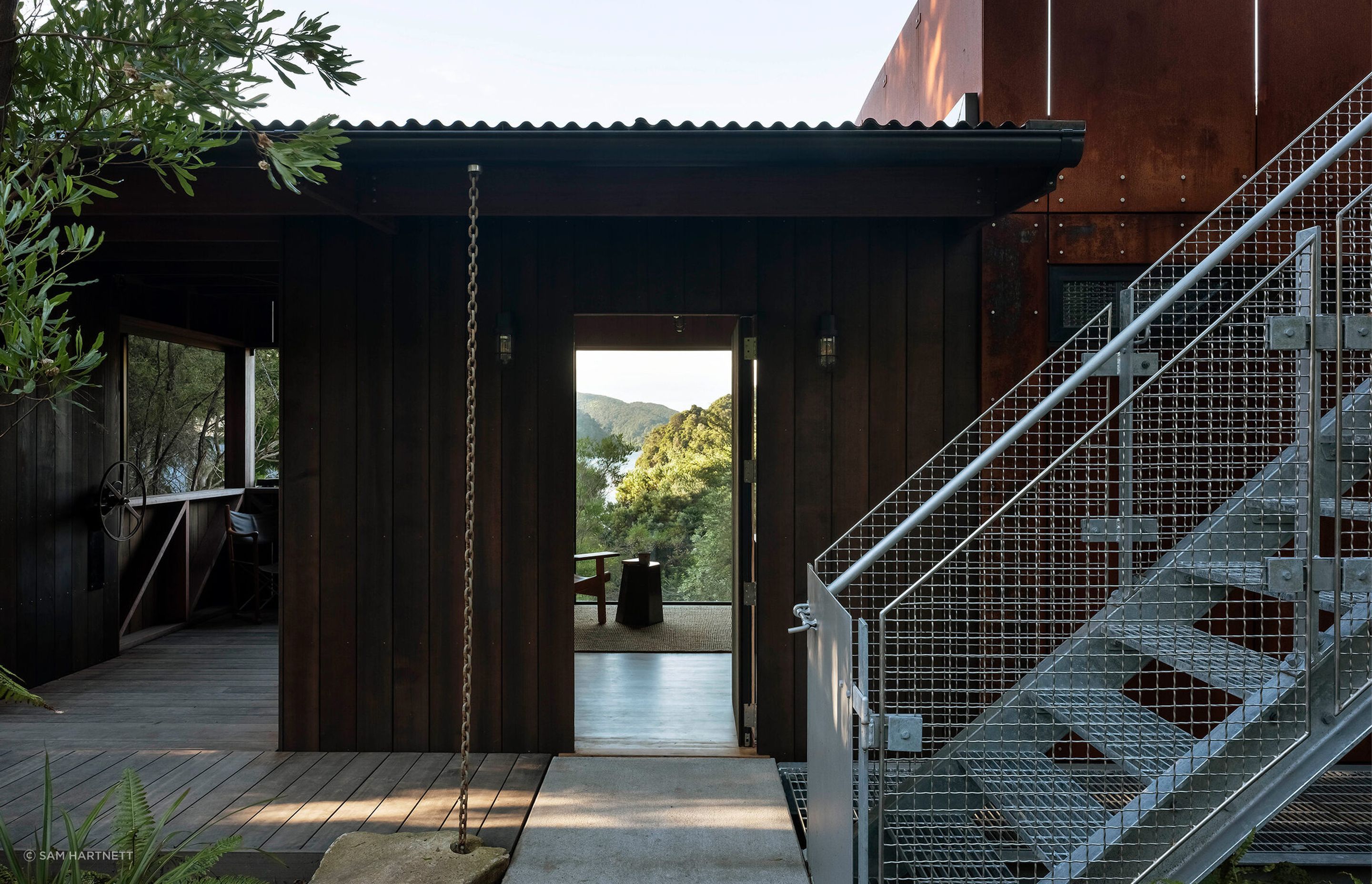 A metal staircase climbs to a treetop roof deck where the Cor-Ten cladding extends into a cantilevered balustrade.