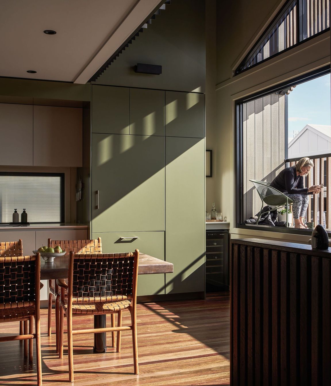 The palette features soft hues of sage and pale greens paired with natural timbers. To the right of the kitchen, a small balcony overlooks the street, capturing the morning and midday sun as it’s filtered through the timber batten screen.
