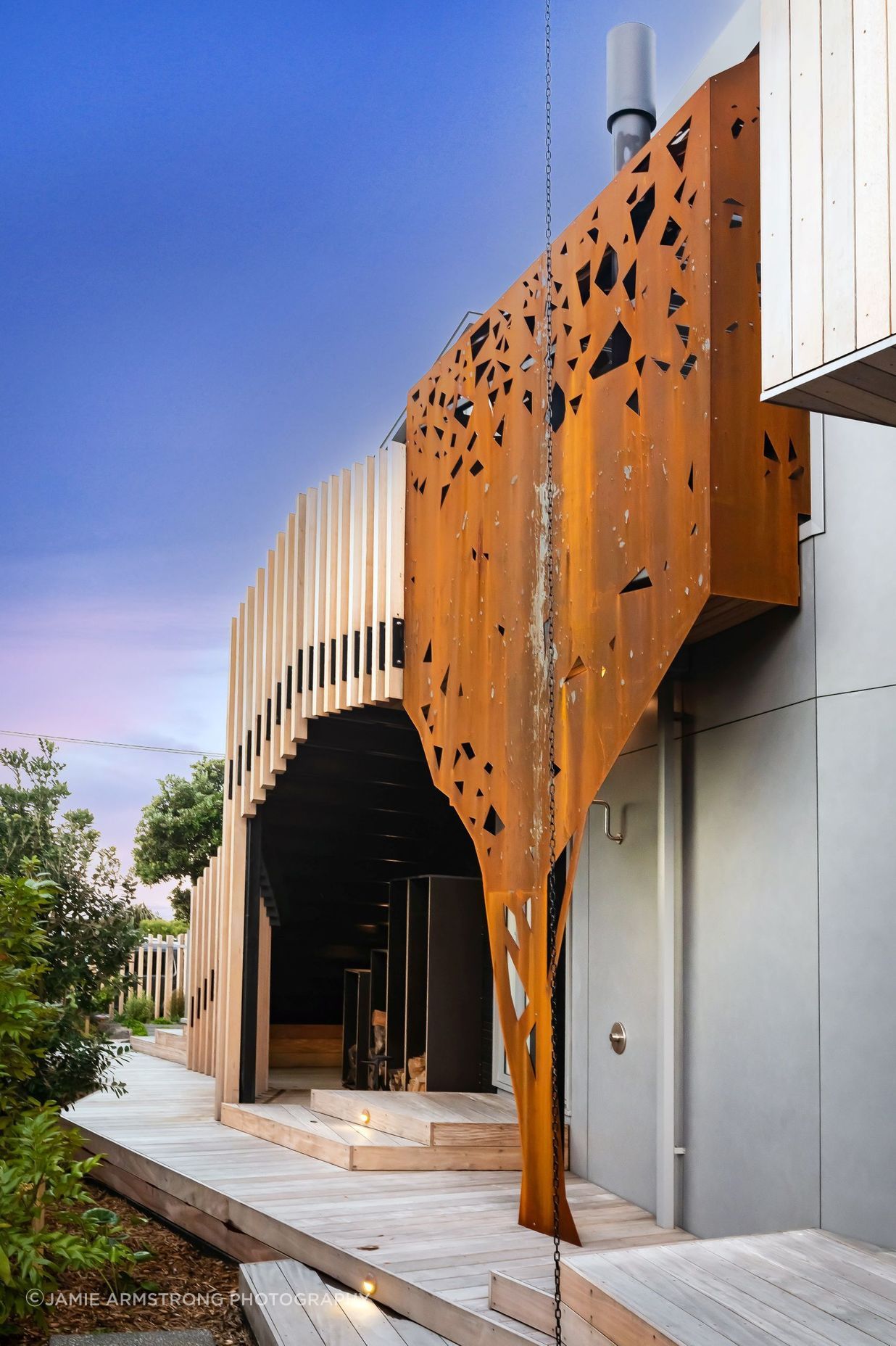 Wrapping around the outdoor fireplace, corten steel has been formed to mimic the silhouette of a pōhutukawa tree across the road from the house.