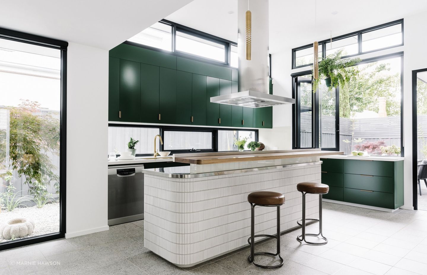 The island bench is clad in kit-kat tiles with brass light fittings above fitting with the rest of the home's mid-century modern details. A timber bar bench invites guests to sit, relax, and join in conversation while food preparation is underway.