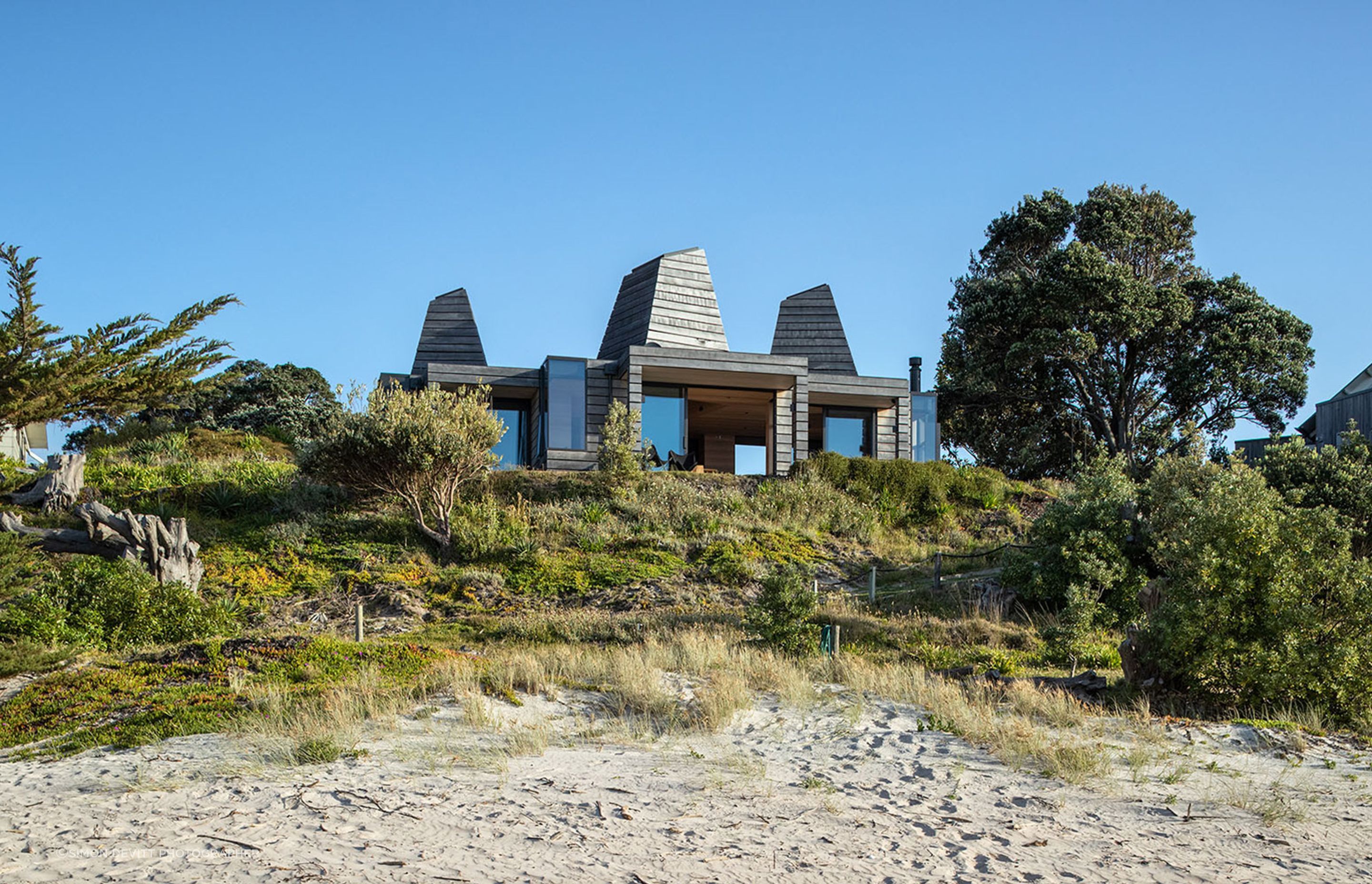 The site rests on the crest of a sand dune, overlooking the ocean and the shadowed curves of distant hills.