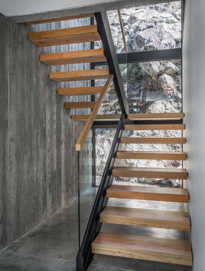 Double-height glazing exposes the rugged cliff face upon entry, forming a textural, organic backdrop to the timber staircase.