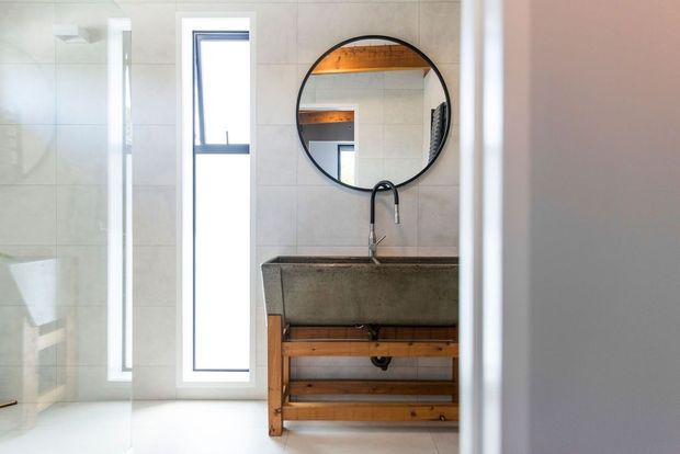 A bright, minimalist bathroom viewed through a doorway, featuring a concrete vessel basin on an open timber vanity, a black wall-mounted faucet, round black-framed mirror, pale tiled walls, white flooring, and narrow vertical windows. The mirror reflects timber ceiling or shelving details and additional glazing.