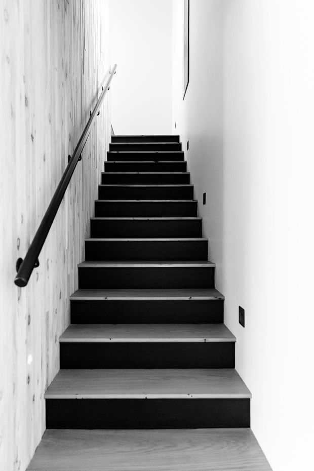 A narrow, minimalist interior stairway viewed from the bottom, with dark stair risers and light timber treads ascending between a pale textured timber wall and a smooth white wall. A simple dark handrail runs along the left wall, with sparse dark wall fixtures and strong high-contrast lighting.