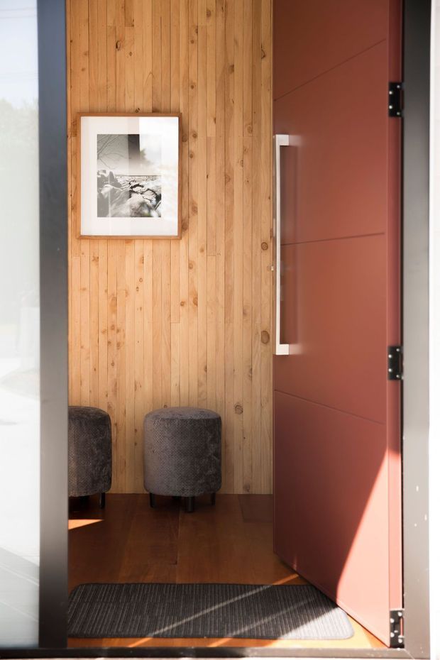 A warm, compact entryway featuring a vertical natural timber-panelled wall, framed monochrome artwork, two dark upholstered stools, and a large reddish-brown hinged door with a long vertical pull handle. Dark wood flooring and a charcoal textured entry mat complete the space, with strong sunlight entering from the side.