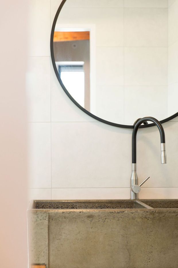 Close-up of a contemporary bathroom vanity featuring a large round black-framed mirror, a wall-mounted or integrated concrete basin with speckled stone-like surface, and a tall black-and-chrome gooseneck tap. Large pale rectangular wall tiles create a clean, restrained backdrop.